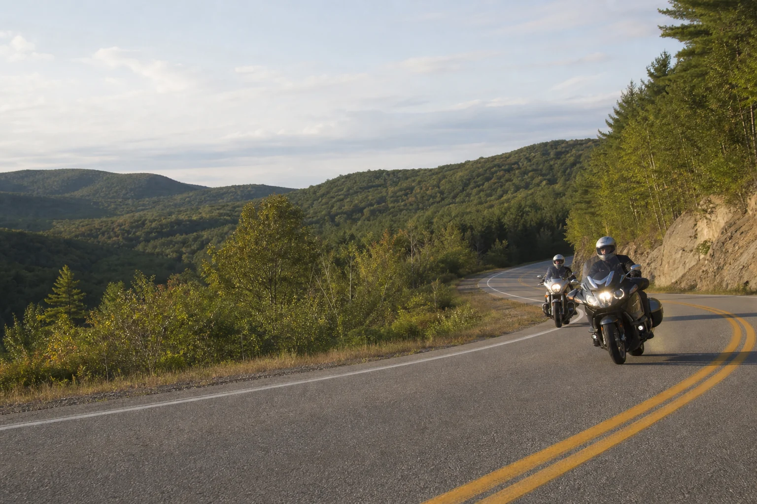 Two touring motorcyclists riding a winding road through wooded hills
